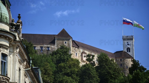 Mighty castle with two flags on a tower nestled in green trees against a clear sky, Ljubljana, Slovenia