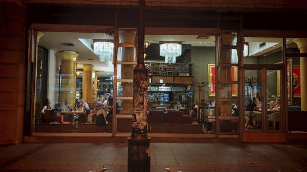 Interior view of a café with glass façade and visible guests at night, Skopje, North Macedonia