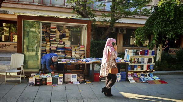Book stand on a street surrounded by people and green trees, idyllic street scene with woman talking on the phone, Pristina, Kosovo