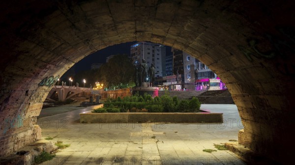 View through a stone archway of an illuminated square with monuments at night, Skopje, North Macedonia