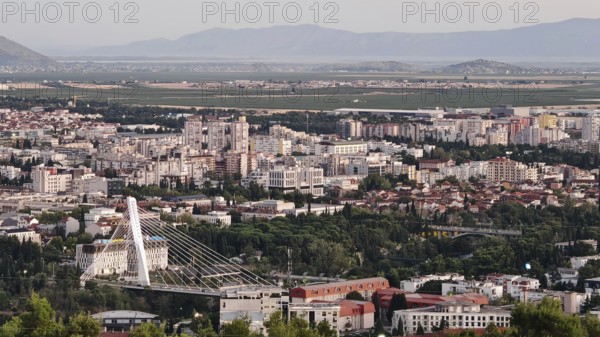 A city surrounded by nature with trees in the foreground and mountains in the background, illuminated by a warm sunset, looking out from a hill on the outskirts of Podgorica, Montenegro