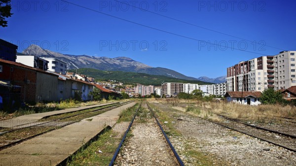 Overgrown train tracks run through a town at the foot of mountains on a sunny day, Kosovo