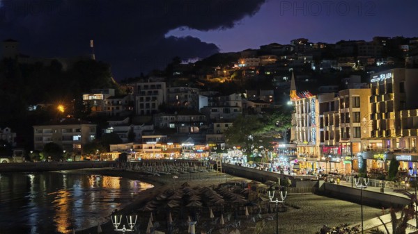 A nighttime beach view with illuminated buildings and dark skies in a lively coastal town, Ulcinj Promenade, Montenegro