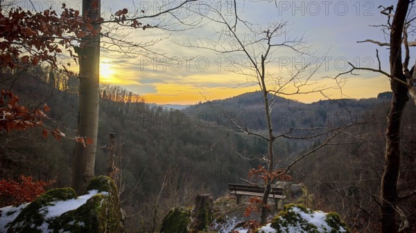 A sunset with a bench with a view over a forest with trees and rocks with snow, Franconian Forest nature park Park