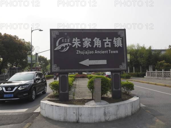 A road sign points to the ancient city of Zhujiajiao surrounded by trees and a road with cars, Shanghai, China