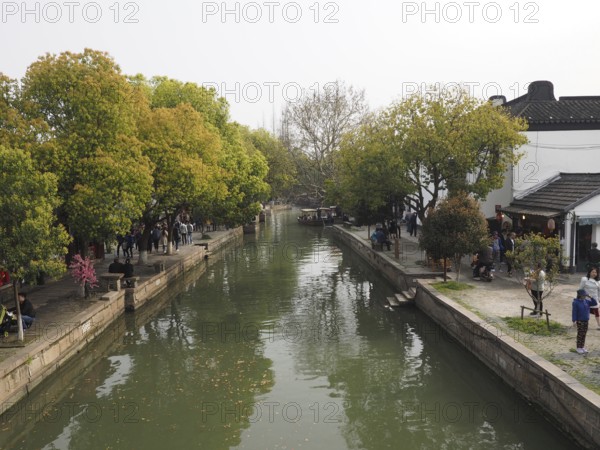 A quiet canal in Zhujiajiao with trees, footpaths and traditional buildings, visited by people, Zhujiajiao Ancient Town, Shanghai, China