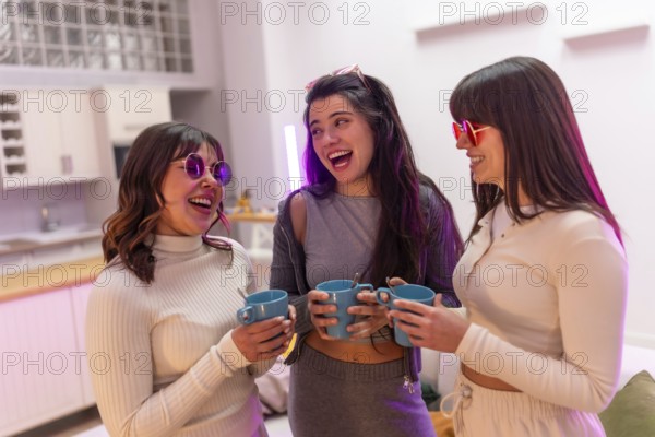 Three smiling young women friends wearing stylish sunglasses and casual clothes sharing a happy moment together, holding mugs and laughing during a cheerful indoor gathering