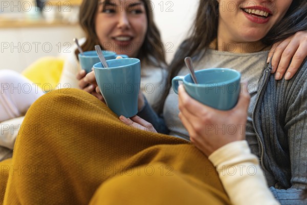Two young women friends enjoying hot drinks and conversation while relaxing on a sofa at home, sharing moments of friendship and comfort under a warm blanket