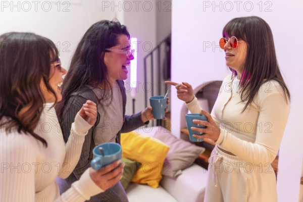 Three young women laughing and chatting over hot drinks in a cozy apartment, enjoying a relaxed evening of friendship, warmth, and casual conversation