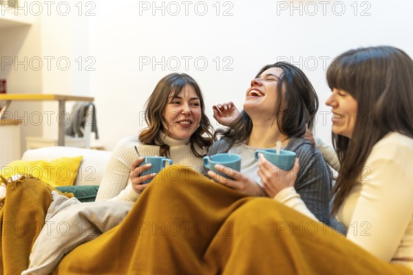 Happy women friends enjoying a cozy moment together at home, laughing and having hot drinks while relaxing on the couch under a warm blanket in an authentic environment