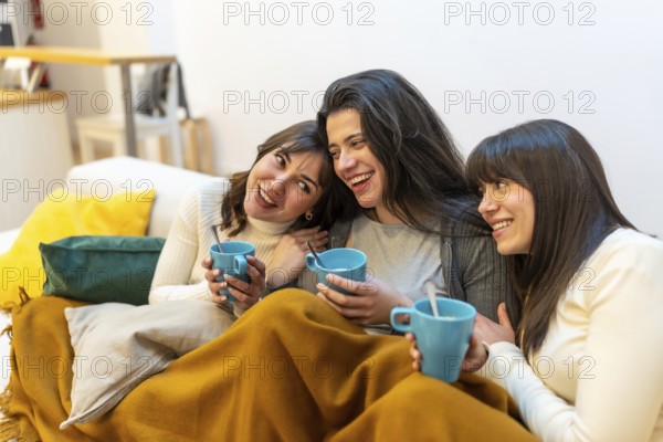 Three young women on a sofa wrapped in a blanket, sharing hot drinks, laughing and chatting in a cozy apartment for a relaxed, intimate moment of friendship and comfort