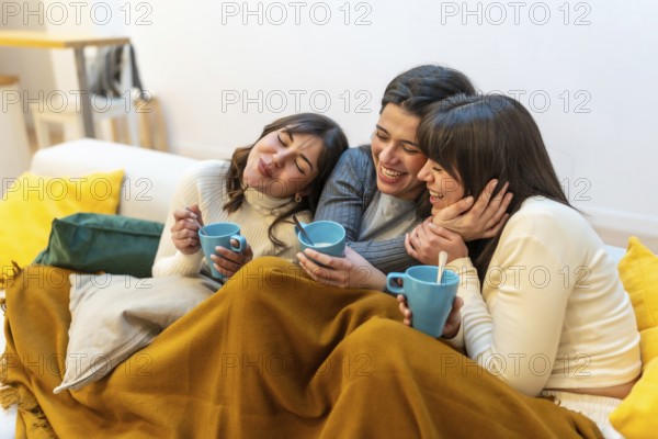 Three young women on a sofa sharing a cozy blanket, laughing and sipping warm drinks while enjoying a relaxed, intimate moment of friendship and comfort at home
