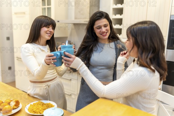 Young friends clinking coffee mugs and laughing in a modern kitchen, enjoying breakfast with cereal and muffins, sharing a warm, casual morning moment together