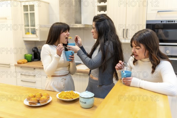 Three smiling young women sharing muffins and cereal, laughing and chatting over breakfast in a bright modern kitchen, relaxed roommates enjoying a casual morning together