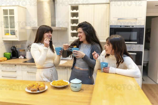 Three smiling young women friends enjoying breakfast together, eating cereal and muffins, drinking from cups, and sharing laughter in a bright modern kitchen