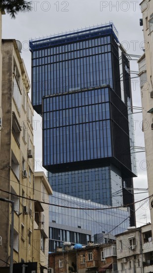 Modern high-rise building with glass façade between older buildings in an urban environment under cloudy sky, Tirana, Albania