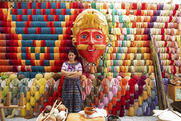 Maya woman, 42 years old, next to a mask of a Moro or Moor for the traditional mask dance, behind a wall full of colorful cotton, traditional crafts in a woman's cooperative, San Juan la Laguna, Atitlán, Sololá Department, Guatemala