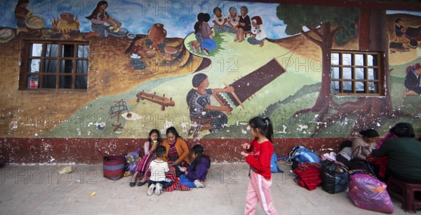 Mayan families in traditional clothes squat in front of a mural at the market in Chichicastenango, Highlands, El Quiché Department, Guatemala