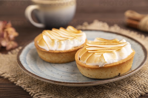 French lemon tart with meringue on brown wooden background, cup of coffee, linen textile, side view, close up, selective focus