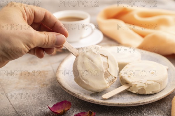 ?hocolate Ice cream in white glaze, cup of coffee, with hand on brown concrete background and orange textile, side view, close up, selective focus, minimalism