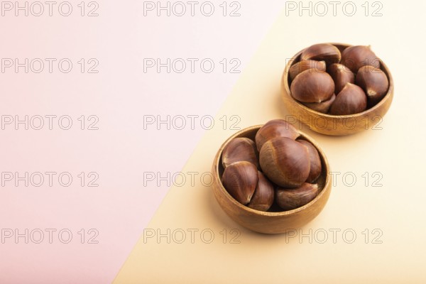 Wooden bowl with raw edible ?hestnuts on pink and orange paper pastel background, side view, copy space, minimalism