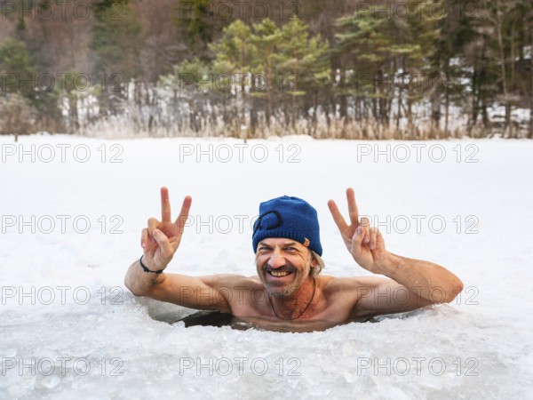 Man with cap, 55, ice bathing in an ice hole makes the Victory sign with both hands, Thumsee, Bad Reichenhall, Berchtesgadener Land, Upper Bavaria, Bavaria, Germany