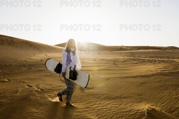 Young woman with snowboard in the sand dunes, near Merzouga, Meknès-Tafilalet region, Erg Chebbi, northern Sahara, Morocco