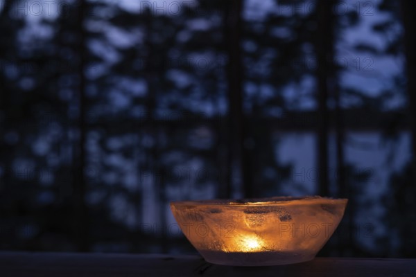 Ice bowl illuminated by burning tea light stands on balcony balustrade, forest at the onset of twilight, winter, Finland
