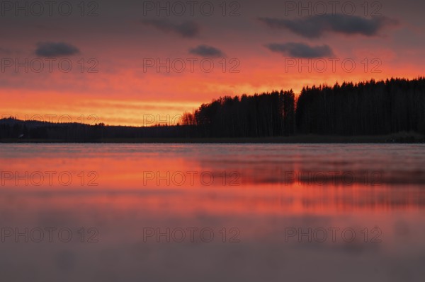 A calm sunset sky with orange-red-yellow hues over a quiet lake and dark forest, Finland