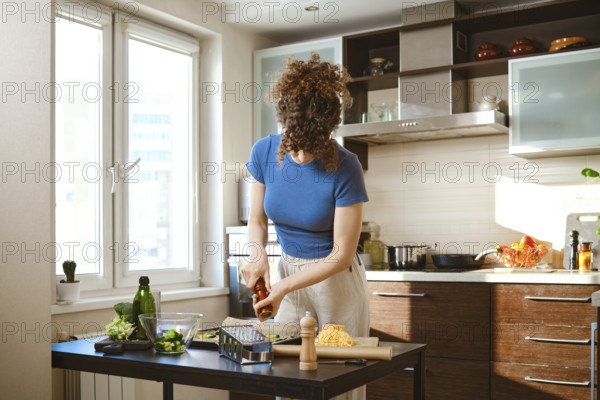 A person prepares smashed broccoli with cheese in bright kitchen. Woman sprinkles pepper over the food adding spice and flavour. Sunlight brightens the space