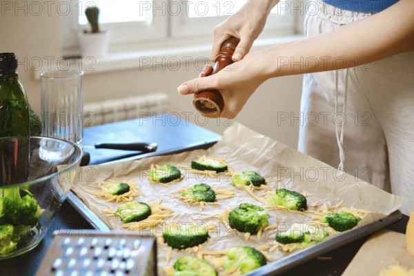Woman sprinkles pepper over broccoli on a baking tray covered with cheese in a kitchen. The scene shows the preparation of the dish. Ingredients and tools are arranged on the counter