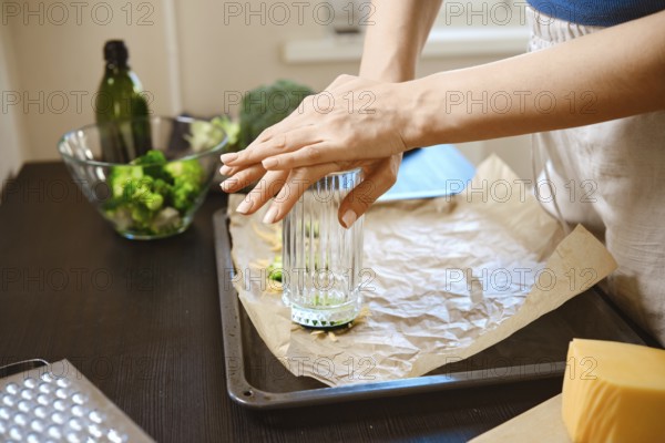 Broccoli is being smashed using a glass in a kitchen. The chef is focused on breaking down the vegetable on a baking sheet. Nearby, cheese and olive oil are ready for cooking