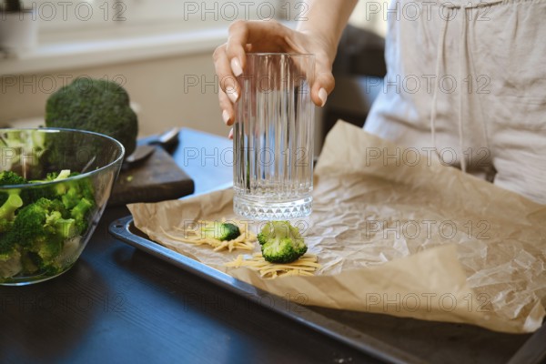 Woman is preparing smashed broccoli with cheese in a kitchen. She is using a glass to smash the broccoli on parchment paper. Fresh broccoli is nearby, showing the steps for a simple dish