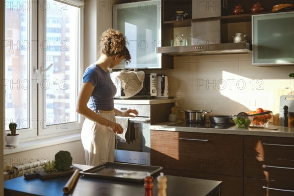 Woman turns on oven to preheat. The sunlight comes through the window. Various ingredients are on the counter, showing a busy cooking moment