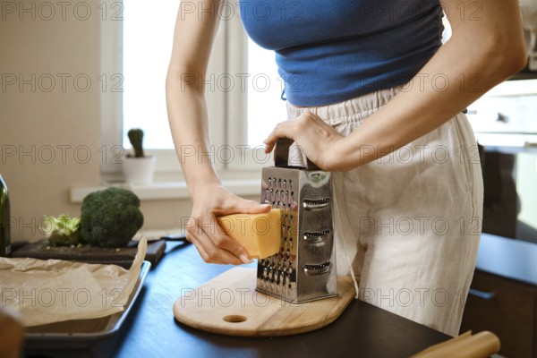 A woman grates cheese over a cutting board while preparing smashed broccoli in a kitchen. The bright sunlight comes through the window. Broccoli and other ingredients are nearby