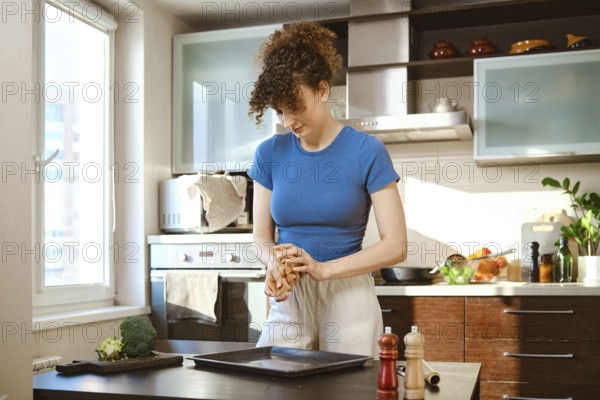 A woman crumples parchment to carefully lay it out on a baking sheet. She stands at a table, pressing the parchment in hands. The kitchen has various cooking tools and fresh ingredients around her