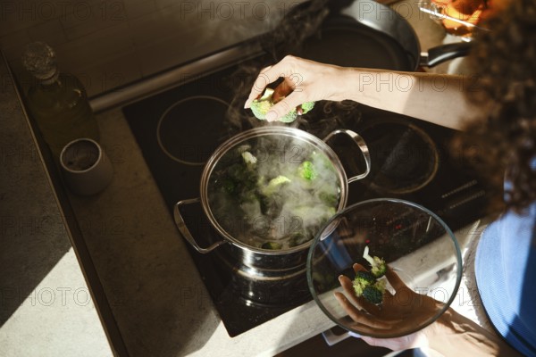 Fresh broccoli is placed into a pot of boiling water on the stove. A woman stands over the pot, ready to cook the vegetables for a tasty dish. Steam rises from the pot as the process begins