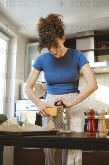 Woman grates cheese in a bright kitchen while preparing smashed broccoli. The scene shows cooking tools on the counter and the person focused on their task