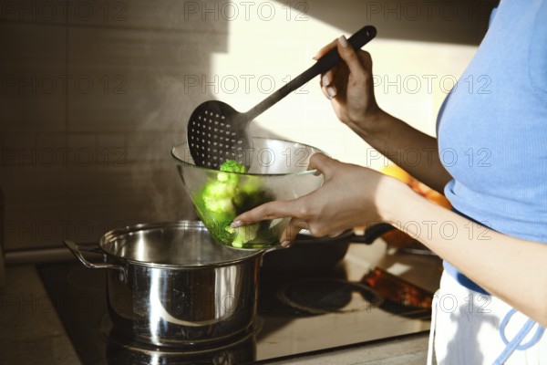 A person shifts steamed broccoli from a pot to a glass bowl using a slotted spoon in a kitchen