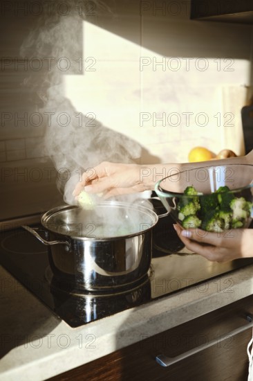 A person is cooking broccoli in a pot on the stove. Steam rises from the pot while another bowl with fresh broccoli is held in the person's hand. The kitchen looks bright with natural light