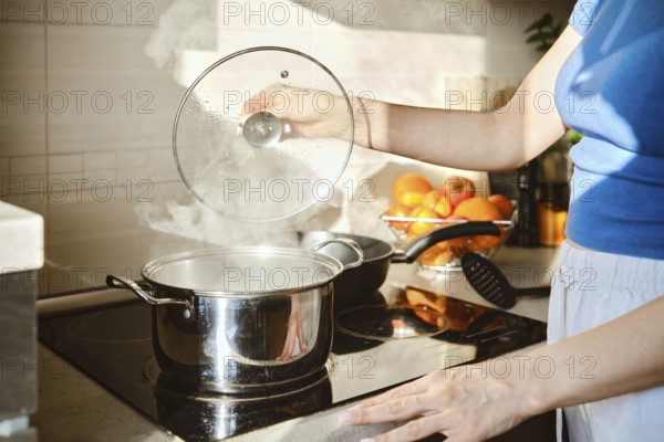 A woman lifts the lid of a saucepan in which water is boiling. Hot steam rises from the pan and is illuminated by the morning sun