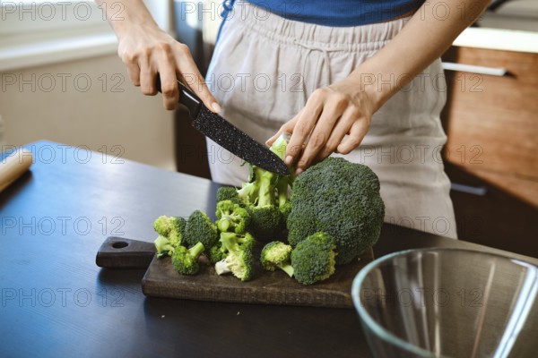 Close-up view of cutting fresh broccoli on a wooden board in a home kitchen. The person uses a knife to chop the broccoli into smaller pieces, preparing for a dish with cheese
