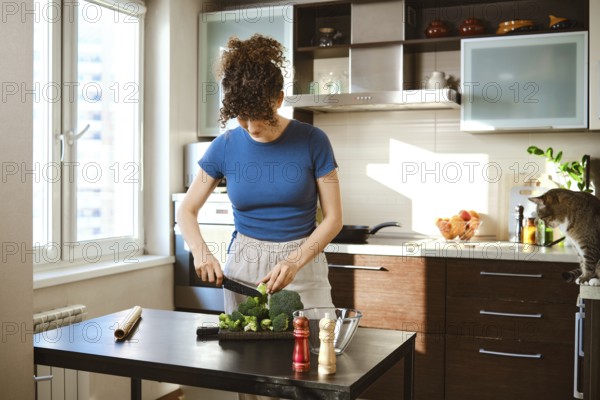 Woman is chopping broccoli on a black cutting board in a bright kitchen. The window shows daylight and a cat sits on the counter. Ingredients for cheese add flavor to the meal