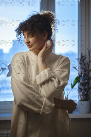 A woman stands softly with her hand on her neck in front of a window. Sunlight fills the room and plants are visible. The setting is bright and shows a calm mood on a typical day