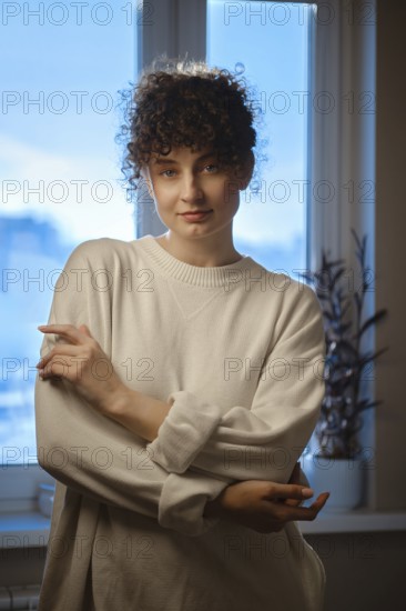 A young woman stands near a window in a dim room. She is wearing a light sweater and looks at the camera. The evening sky outside shows clouds and sunlight coming through the glass