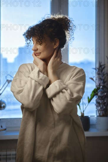 Young woman looks out the window while touching her face in a cozy indoor setting during daytime