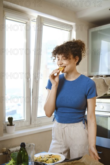 Woman bites smashed broccoli with cheese. The window opens to a city view. Fresh ingredients are on the counter while the person enjoys a bite of food