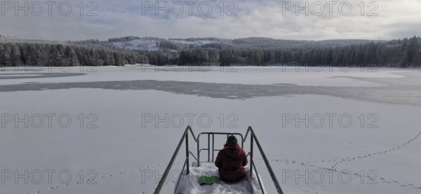 Person sitting on snowy footbridge with a view of the partially frozen lake, Rennsteig, Thuringian Forest