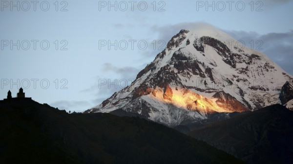Sunbeams illuminate a mountain peak in the evening light with church in the foreground, Gergeti Monastery with Kazbek in the background, Stepantsminda, Georgia
