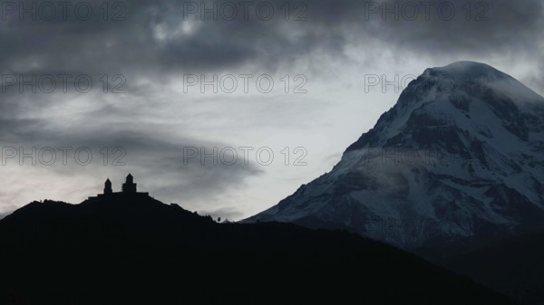 Silhouette of a church in front of a snow-covered mountain at dawn, Gergeti monastery with Kazbek in the background, Stepantsminda, Georgia